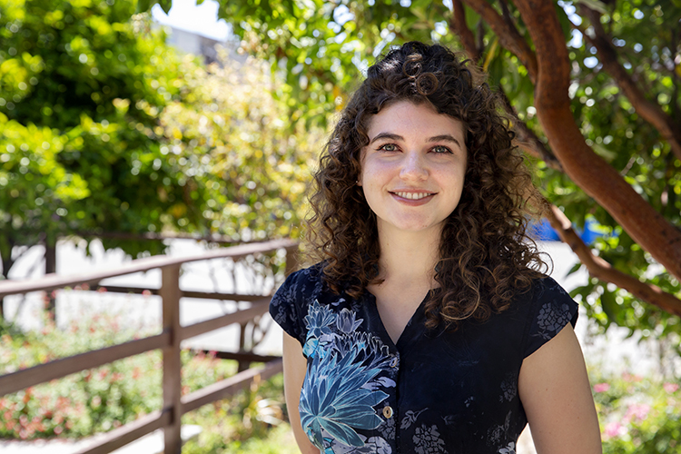 Photo of a young woman smiling into the camera, trees in soft focus behind her