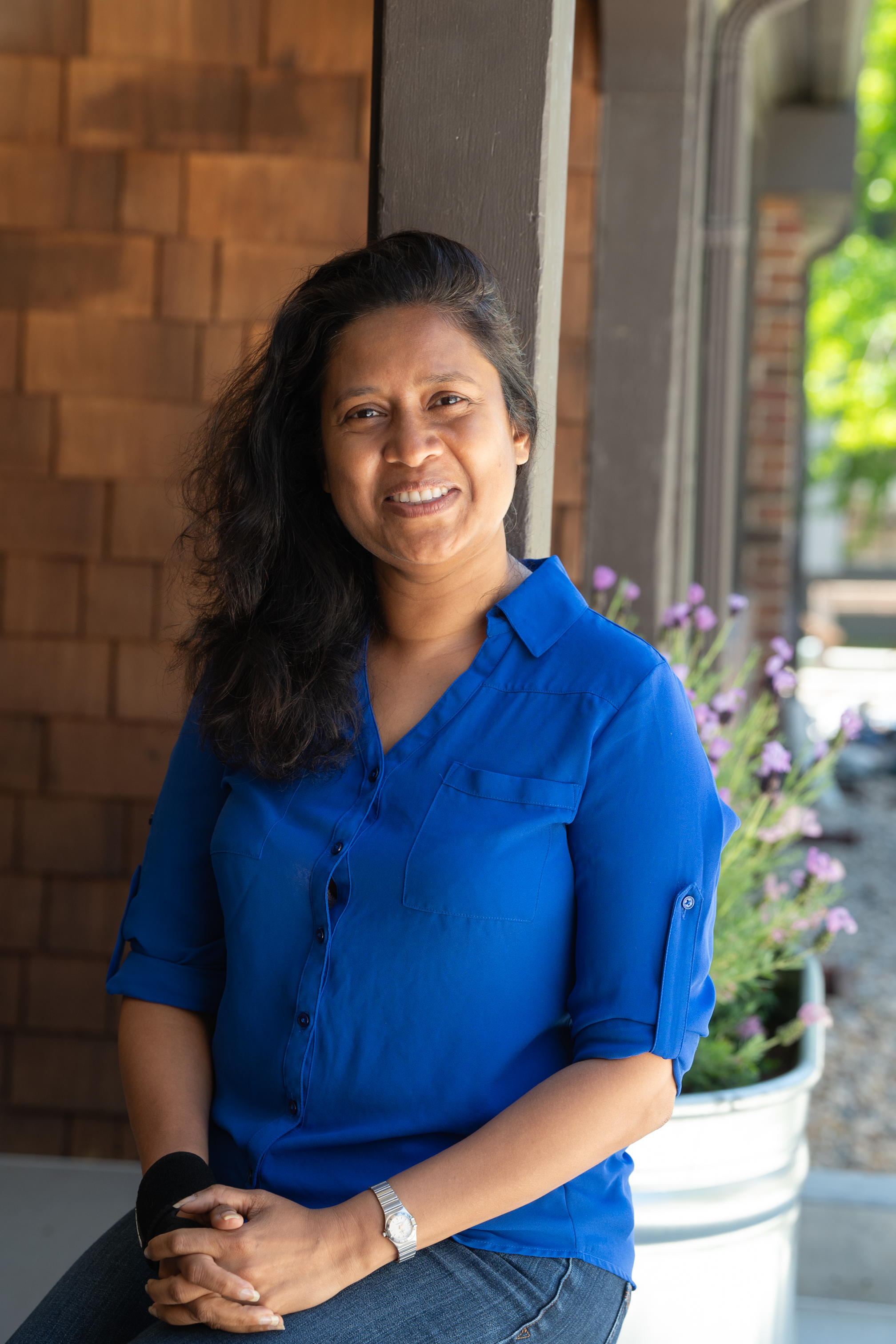 Photo of Sumudu in a blue top, smiling and sitting on a ledge against a wood shingled wall.