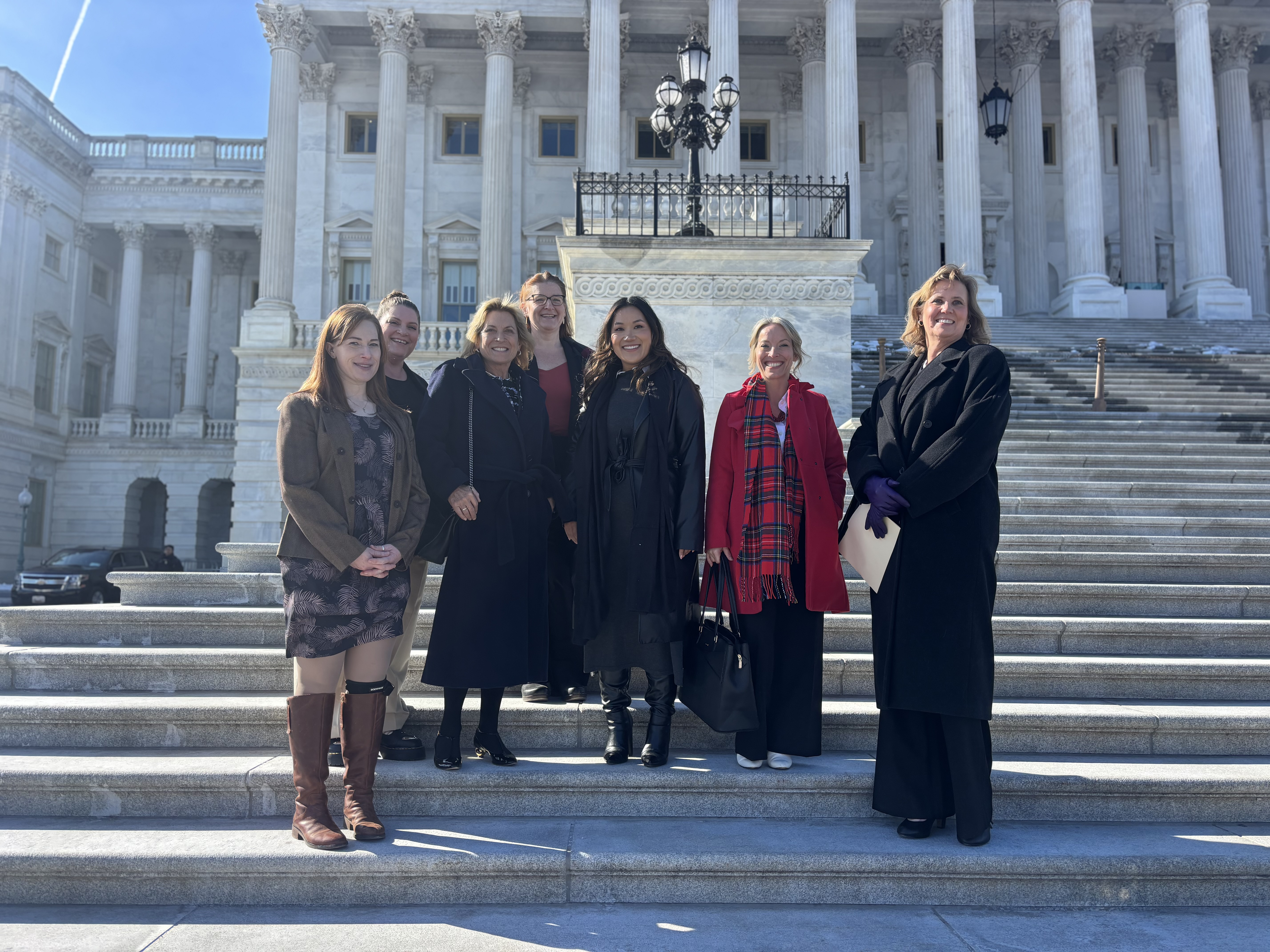 Seven women are standing on the steps of the U.S. Capitol, a neoclassical building that looks like an ancient Roman temple.