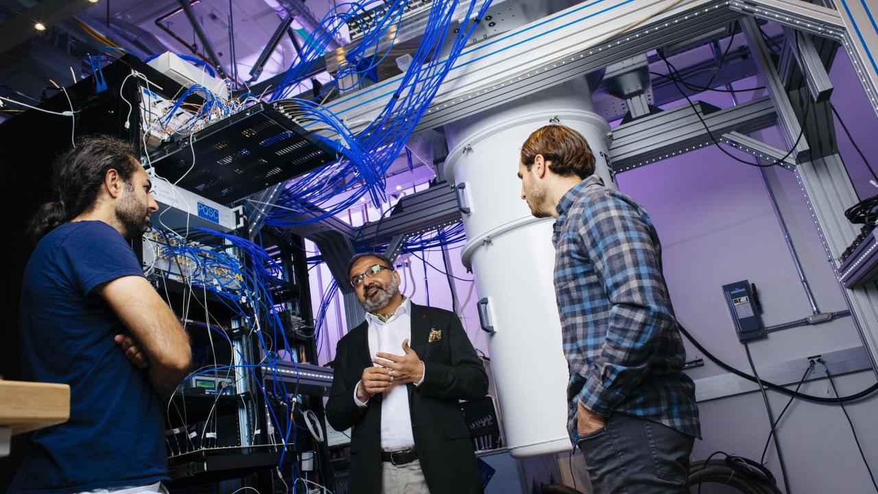 Photo of three men talking in a quantum computing lab awash in a tangle of blue wires and other equipment.