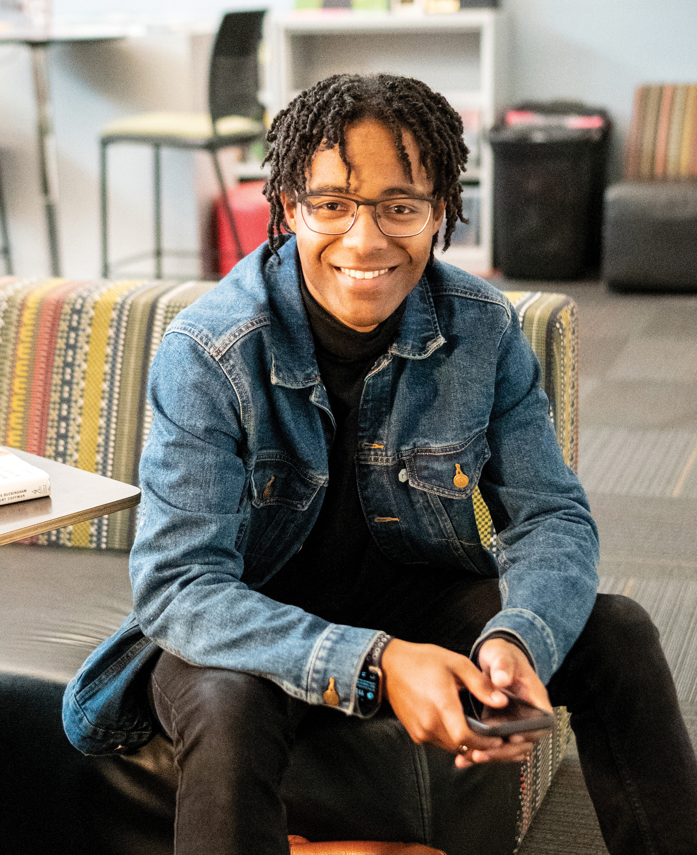 Photo of William sitting on a striped couch, wearing a black top, black pants, and jean jacket