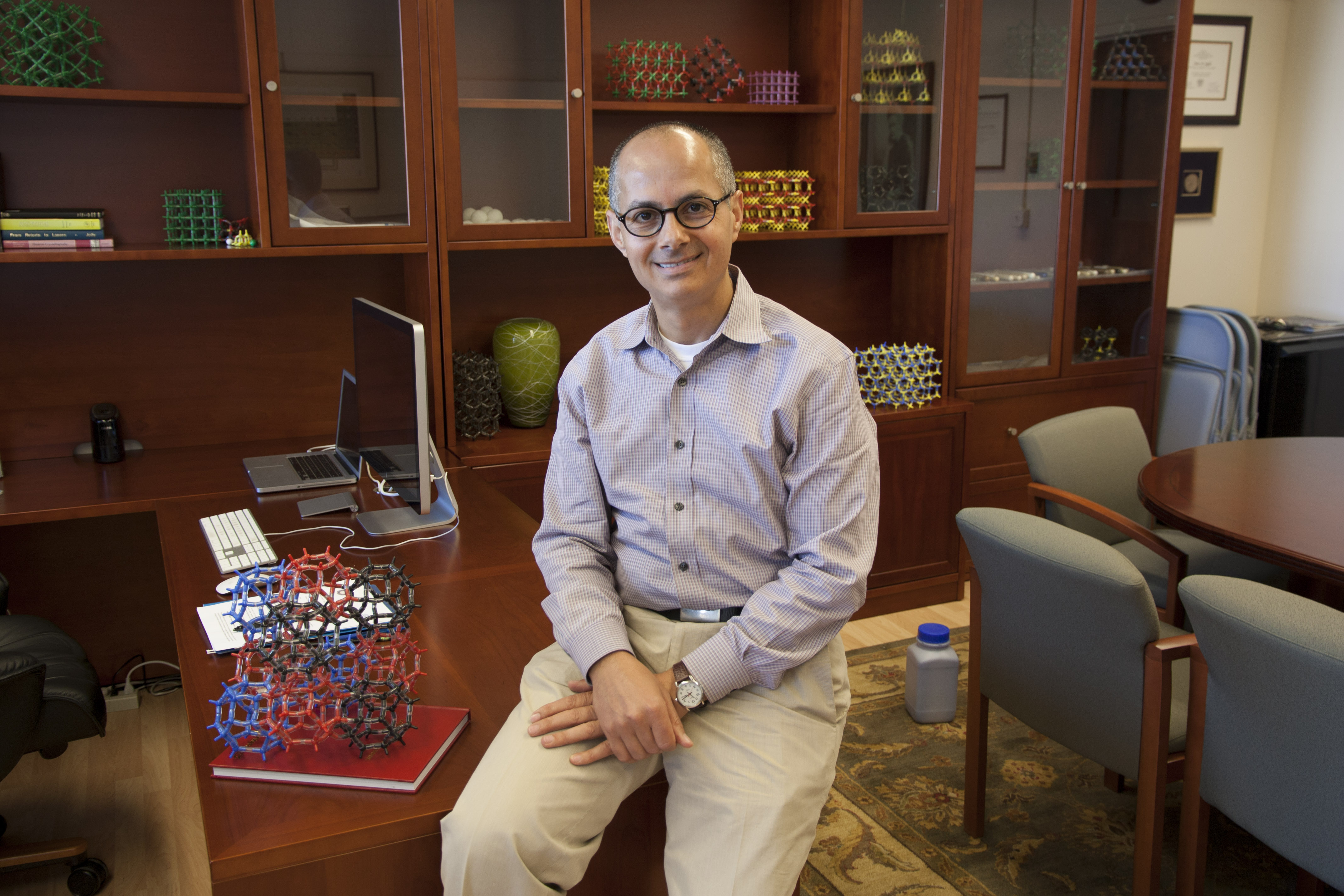 Color photo of a smiling man wearing glasses, sitting on the edge of a desk, surrounded by models of molecules positioned throughout the office.