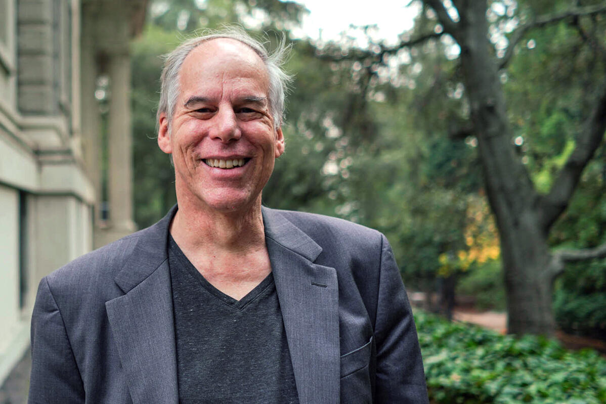 Photo of Alan smiling in a black V-neck shirt and dark gray jacket, with ivy and a tree behind him.