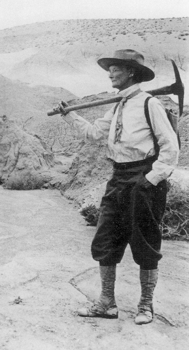 Black and white historic photo of Annie in a Panama hat, white shirt, tie, and riding pants, holding an ax over her shoulder.