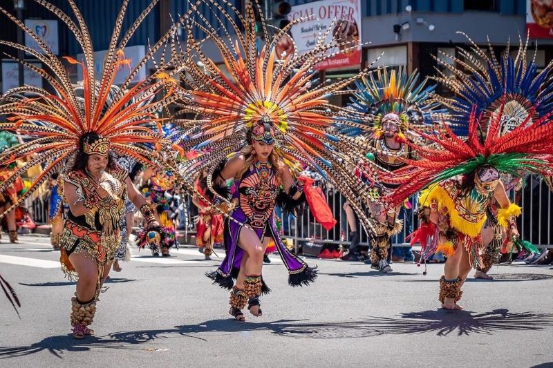 Photo of dancers wearing long, bright feathered headdresses and brightly colored costumes.