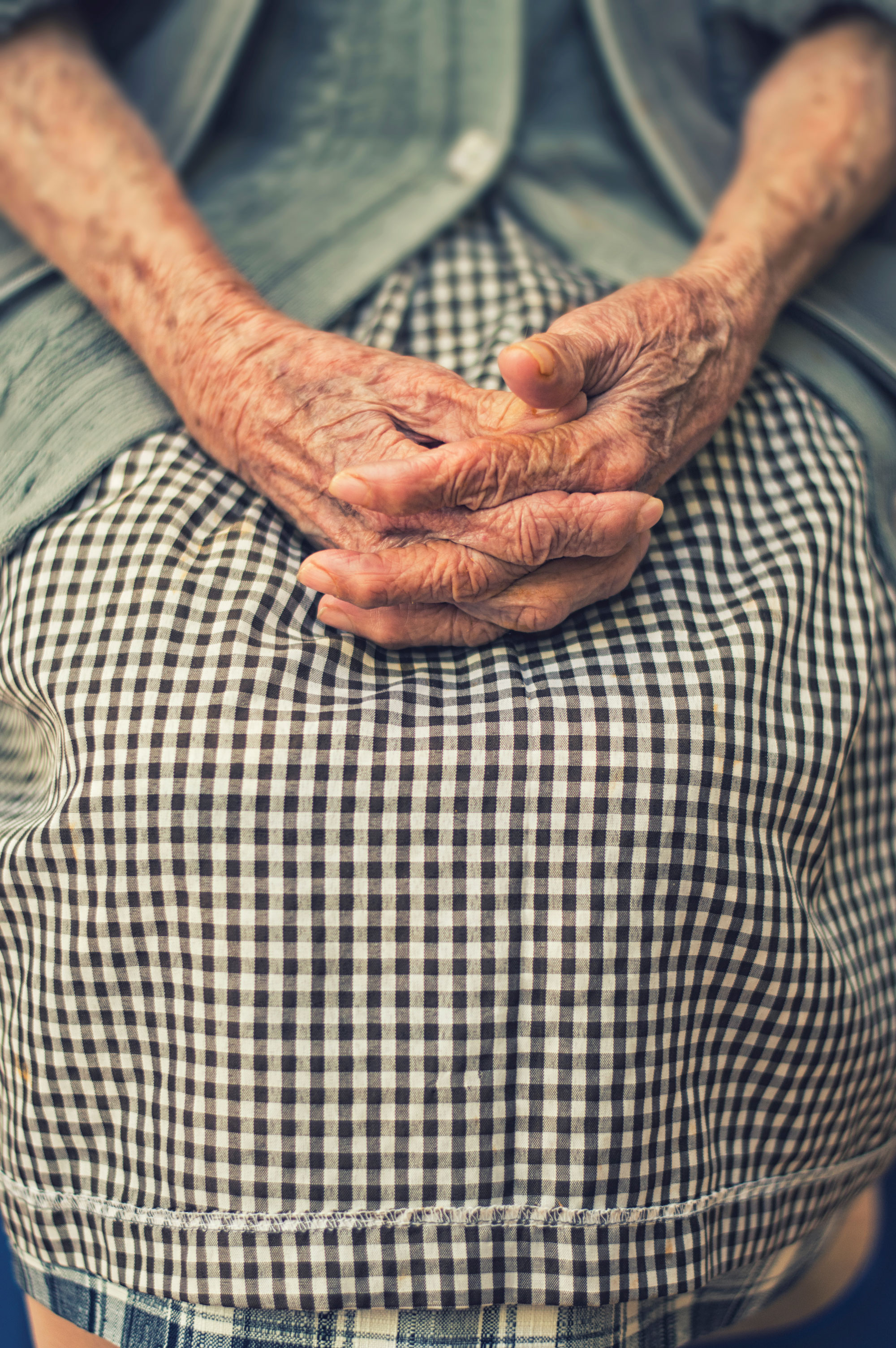 Photo of an elderly woman in a checkered dress and cardigan with her hands clasped on her lap.