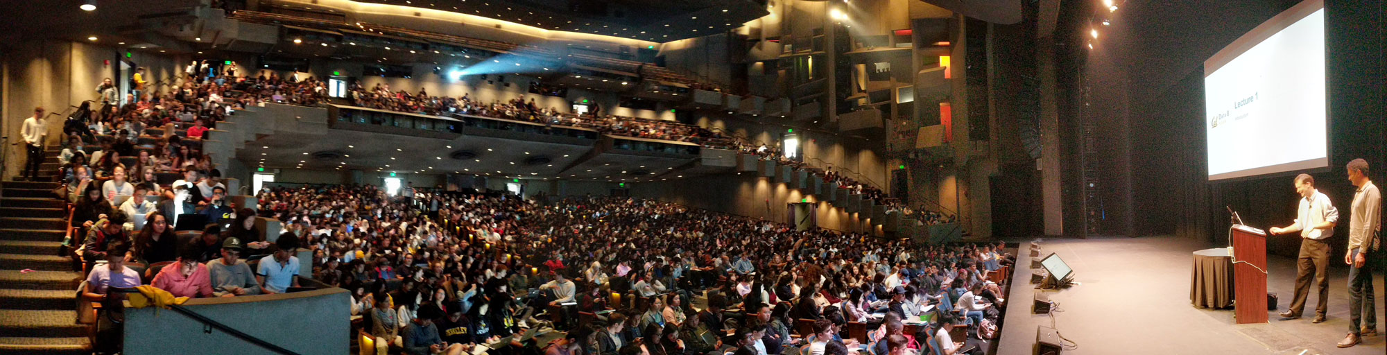 Photo of a very full lecture hall with two men onstage and a slide reading "Lecture 1" behind them