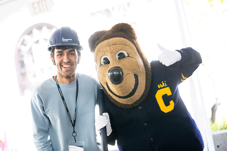 A student in a pale blue sweatshirt and hard hat hugs Oski, who's wearing a navy blue letter sweater and waving.