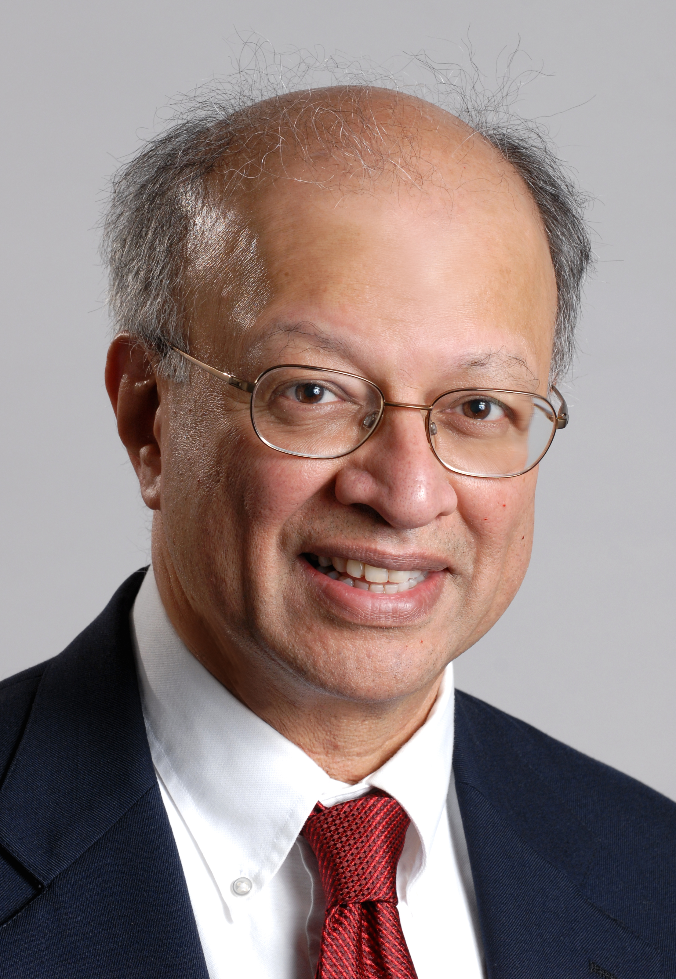 Headshot of Ashok against a neutral gray background, smiling and wearing glasses, a dark jacket, white shirt, and deep red tie.