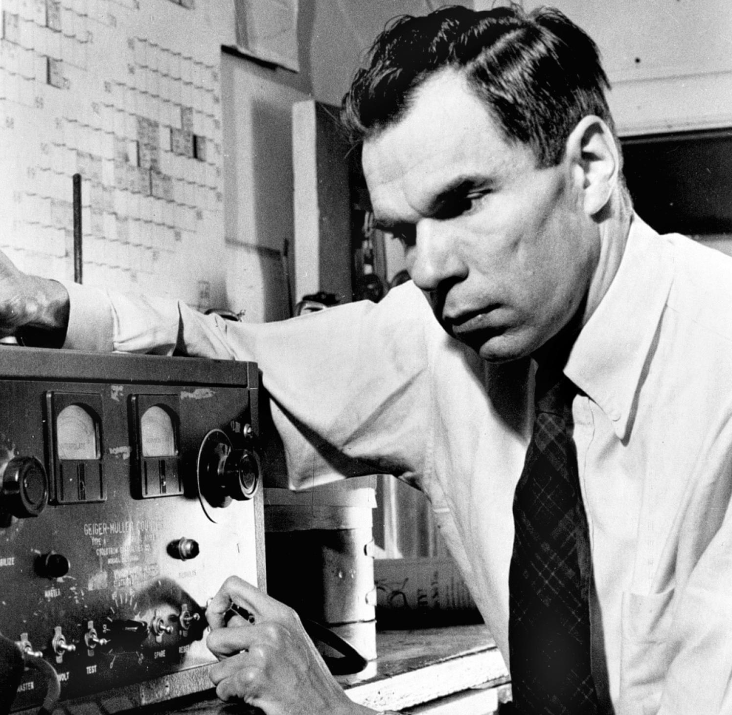 Black and white photo of Glenn in a white shirt and dark tie manipulating dials on a Geiger–Müller counter, an electronic instrument used for detecting and measuring ionizing radiation