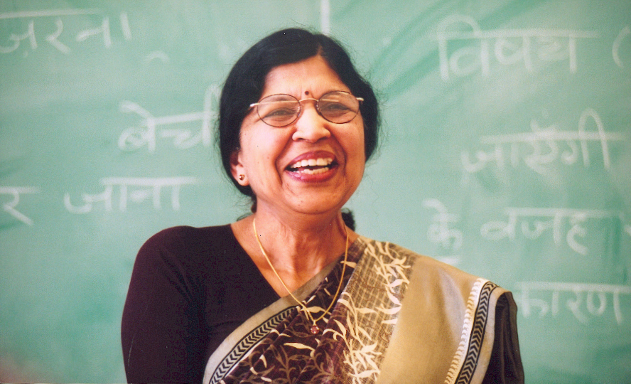 Photo of Usha in front of a chalkboard with Hindi written on it. She is smiling and wearing a sari in brown and purple tones.