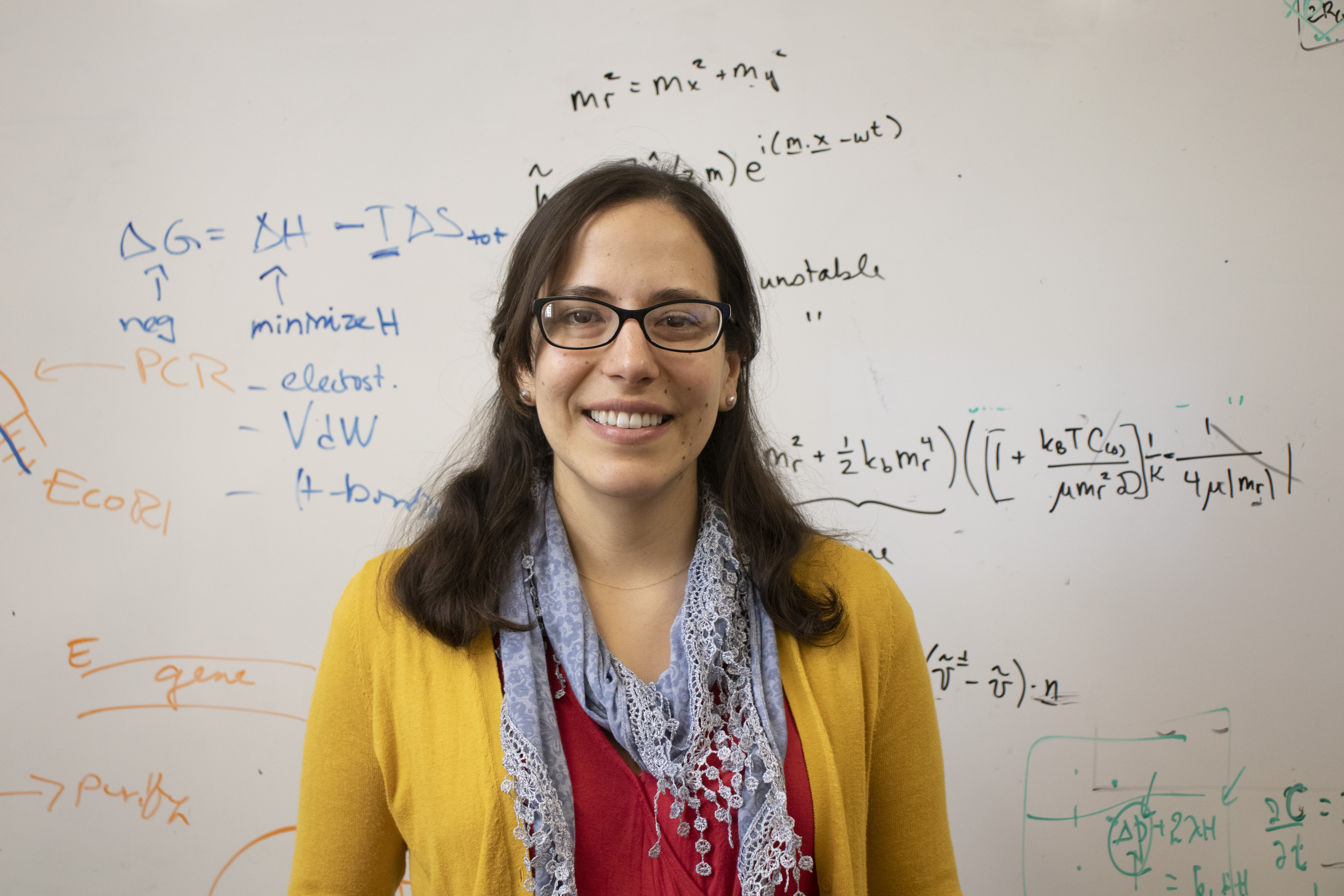 Photo of Markita in a red top, blue scarf, and yellow cardigan, smiling in front of a white board with writing on it.