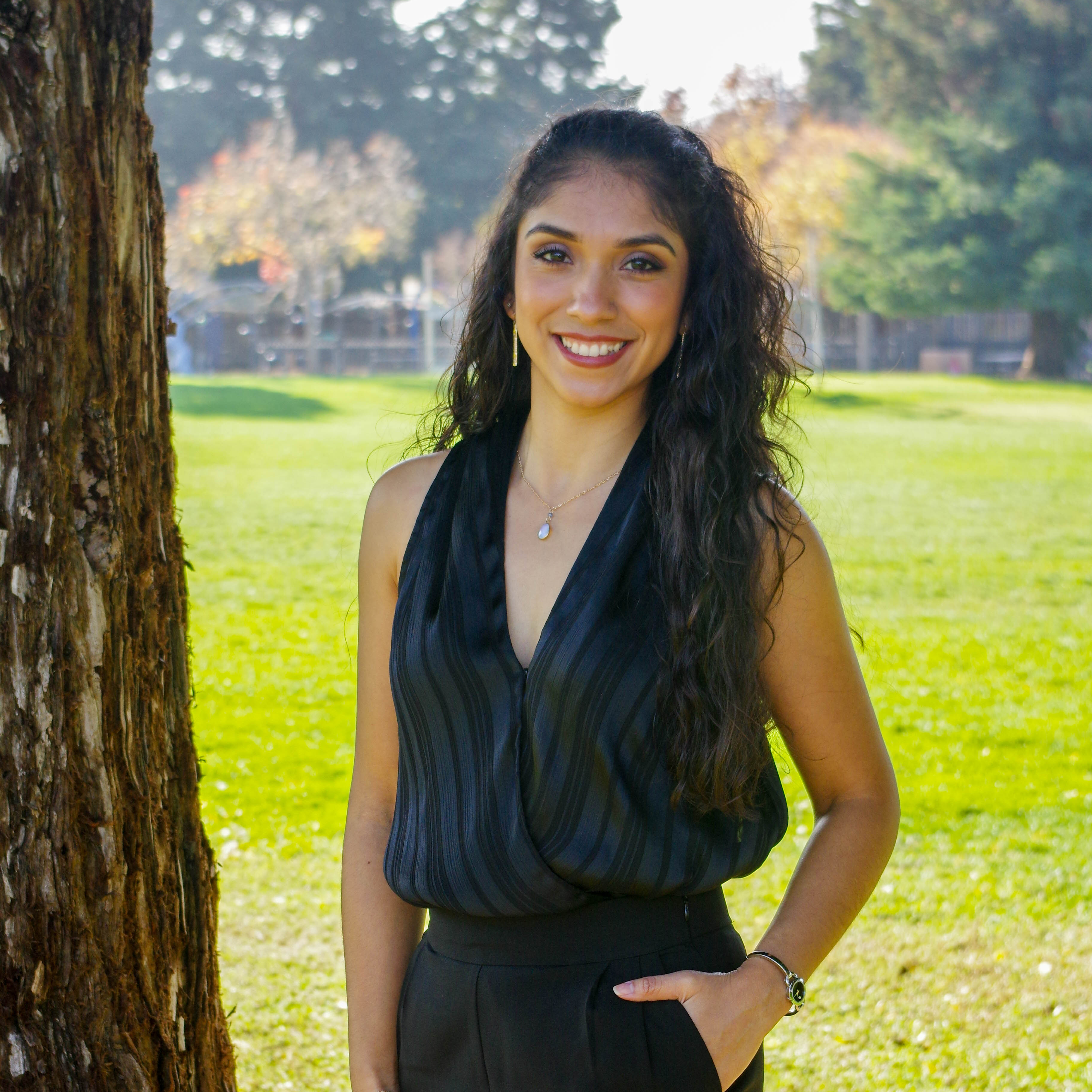 Color photo of Mayra smiling, standing next to a tree on a sunny day, her left hand in her pants pocket.