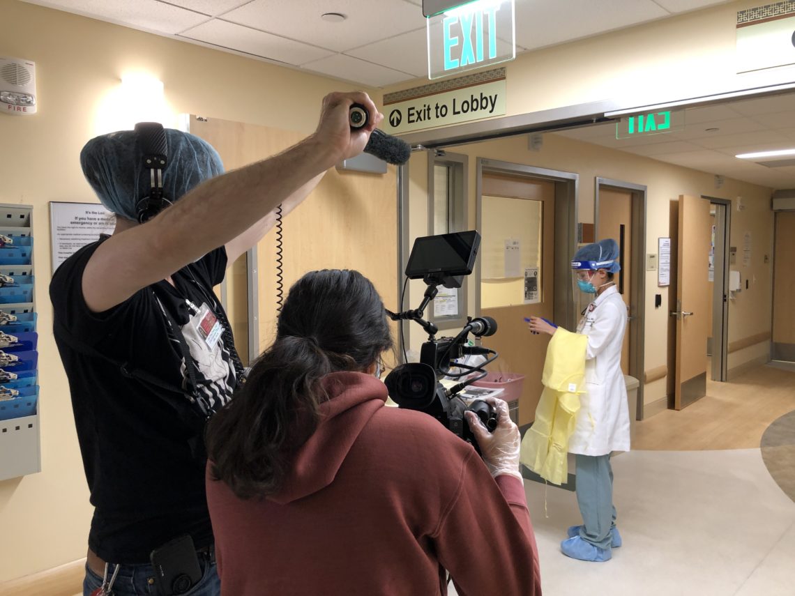 Photo of students, one holding a mic and one holding a camera, filming a doctor in a hospital corridor.