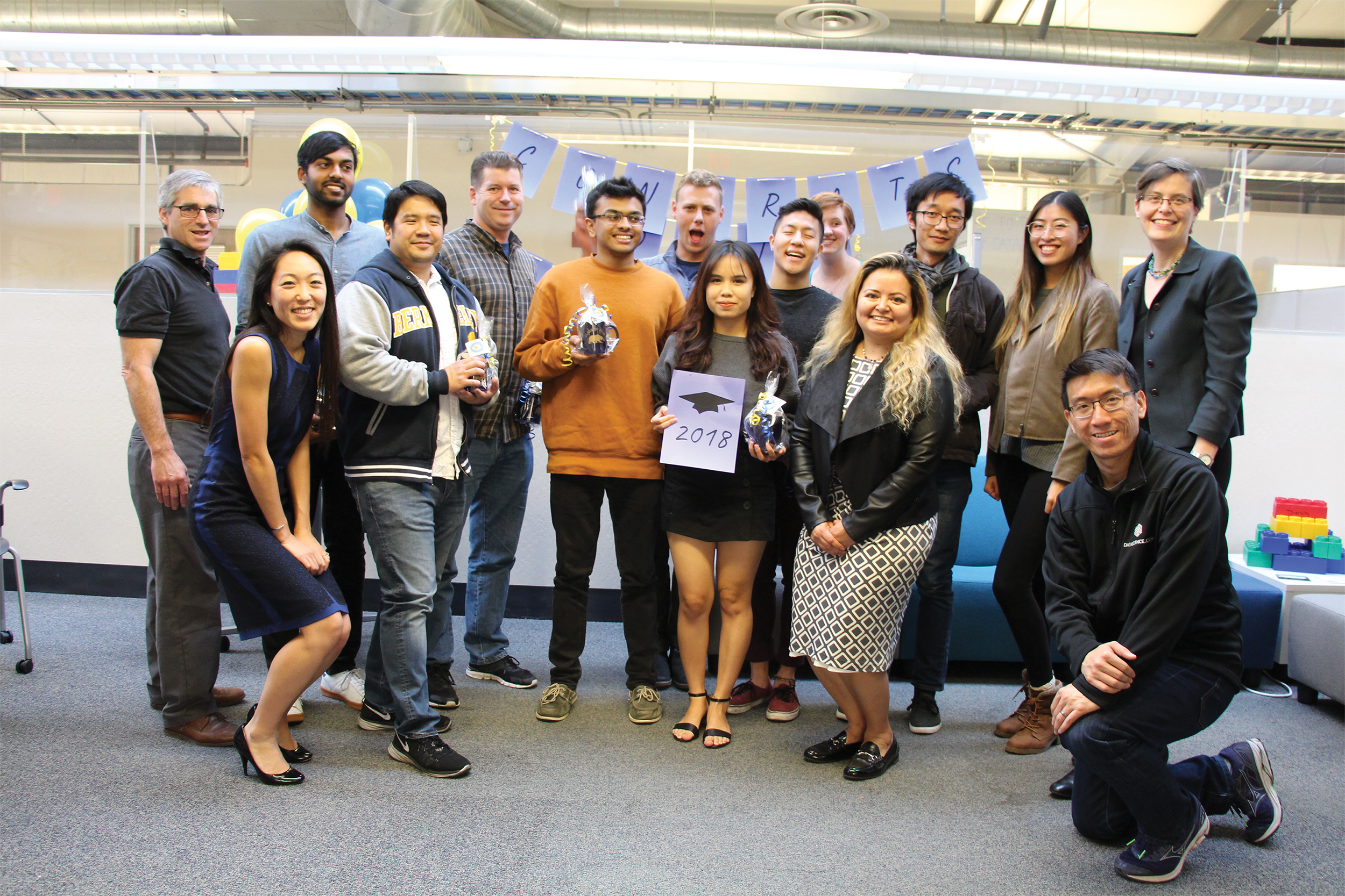 Photo of the graduates and their supporting faculty and staff smiling in front of a homemade 