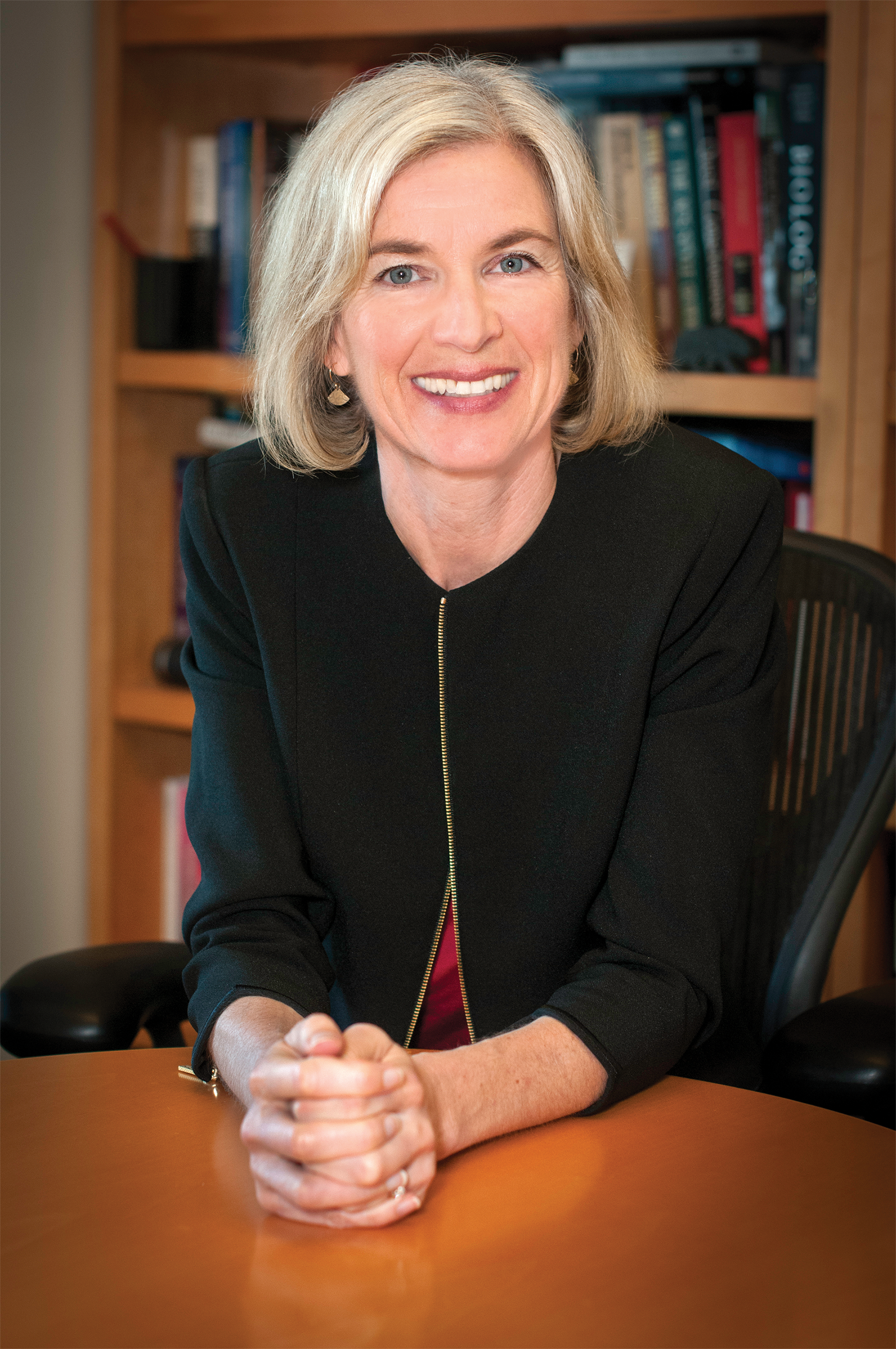 Photo of Jennifer Doudna leaning on a table with a bookcase behind her.