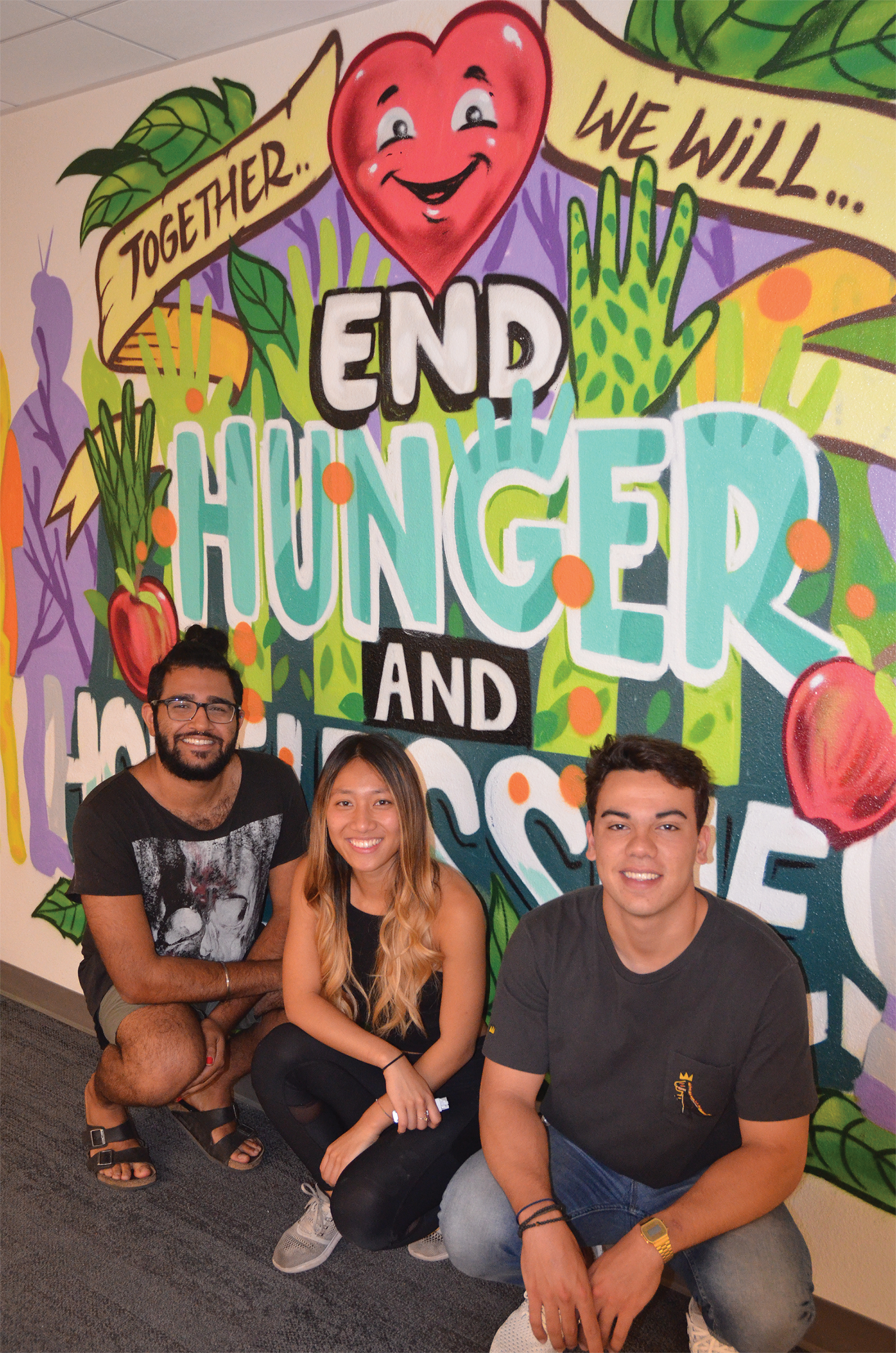 Photo of the staff and student leaders kneeling in front of a brightly painted mural that says, “Together we will end hunger and homelessness.”