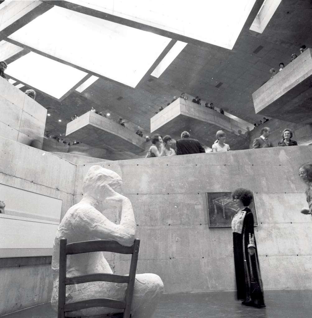 Black and white photo of a woman in a long dress gazing at a large, white sculpture of a man sitting in a chair.