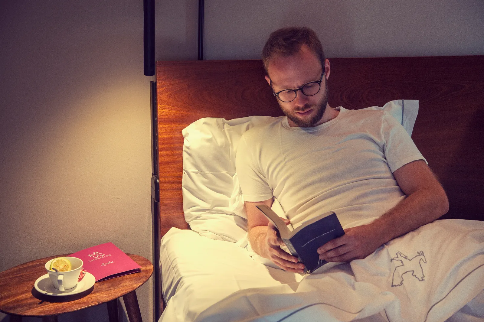 A man relaxes in bed reading a book with a drink on a small table next to him.