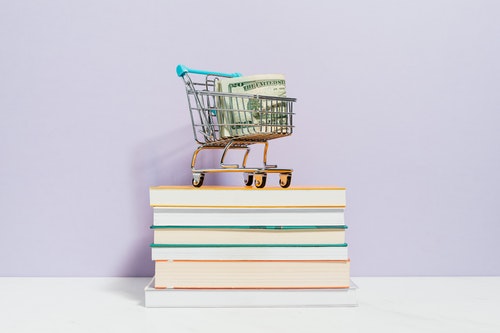 money roll in tiny shopping cart on stack of books lightly colored background