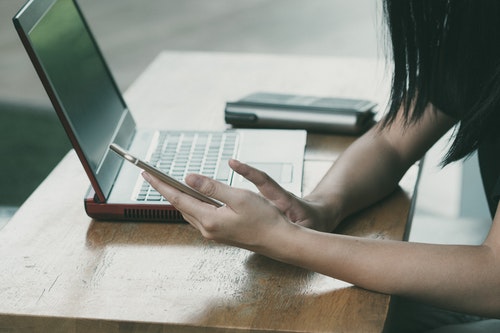 person sitting at table with open laptop with phone in hand