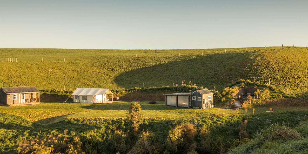 Aerial view of a glamping site in rolling green hills at golden hour, with cabins, a bell tent, and pavilions scattered across a peaceful valley beneath a clear sky.