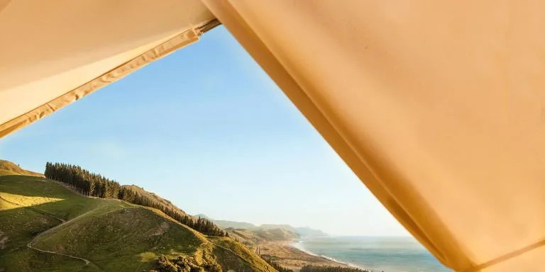 Scenic coastal landscape viewed through a golden tent opening, showing lush green hillside with forest line meeting turquoise ocean waters under clear blue sky