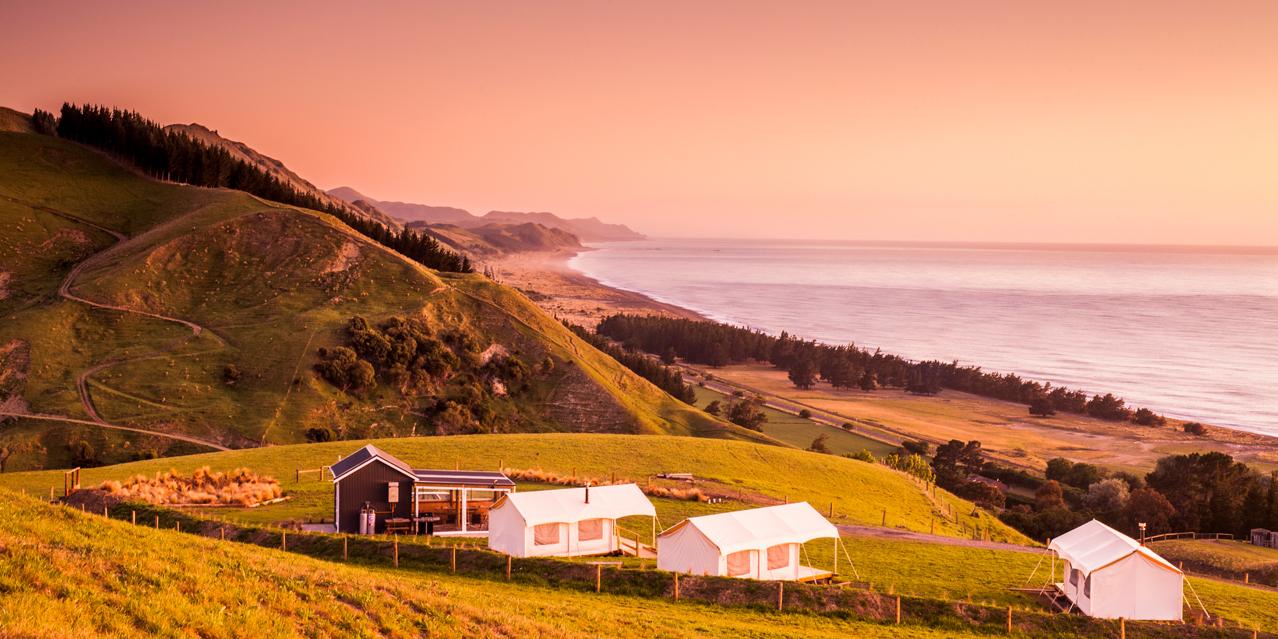 Rolling golden hills with evergreen forests descend toward a misty coastline at sunset, featuring glamping tents nestled on the hillside under a warm peachy sky