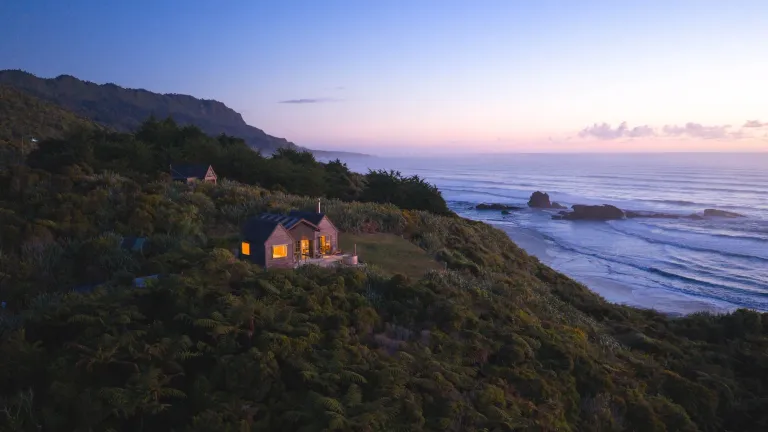 A small cabin with warm lights sits on a bush-covered coastal hillside at dusk, overlooking waves rolling onto a rocky beach beneath a pink and blue sky