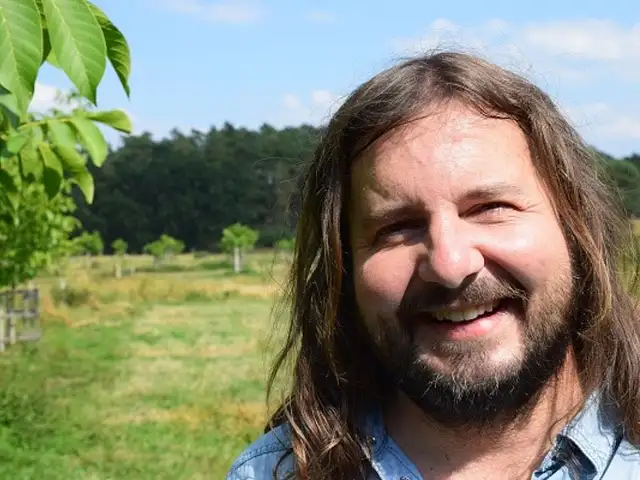 Slowgarden Gründer David (langes, aschblondes Haar und Bart) steht lächelnd auf einer grünen Wiese. Im Hintergrund sind junge Bäume zu sehen.
