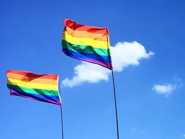 Zwei LQBTQ-Flaggen flattern im Wind. Im Hintergrund ist blauer Himmel mit Sonnenschein und zwei kleine Wolken zu sehen.