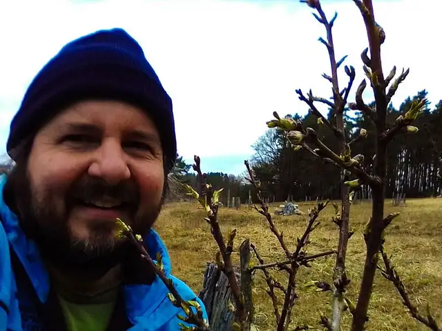 David von Hof Windkind steht auf einem Feld. Vor ihm sind ein paar Zweige zu sehen. Er lächelt in die Kamera. Er trägt einen kurzen Vollbart. Auf dem Kopf hat er eine schwarze Strickmütze und er hat eine blaue Jacke an.