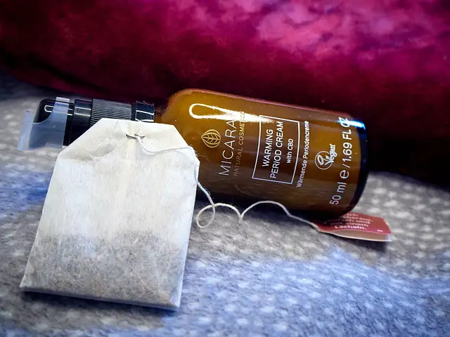 Eine Wärmflasche mit einer lila Samthülle liegt mit einem Teebeutel und einer beigen Verpackung für eine wärmende Periodencreme mit goldener Beschriftung auf einer grauen Decke mit weißen Punkten