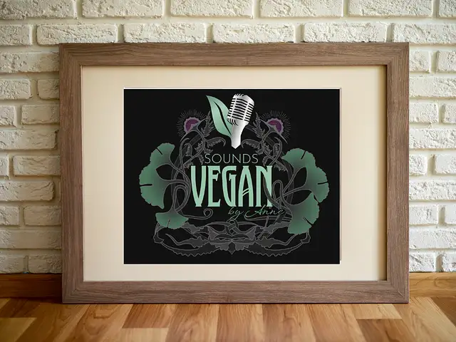 The new Art Nouveau design logo gray with green on black in a picture frame on a raw industrial wall. The lettering reads "Sounds Vegan" and above it is the blog's logo: a microphone with a leaf and two purple thistle flowers.