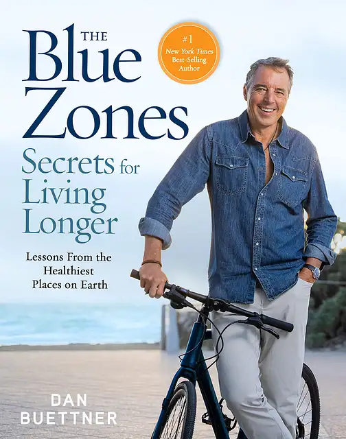 Dan Buettner, ein Mann mit grauen, kurzen Haaren steht in einem blauen Jeanshemd und einer sehr hellen Jeans an eine schwarzes Fahrrad gelehnt und lächelt. Im Hintergrund ist das Blau des Himmels zu erkennen.