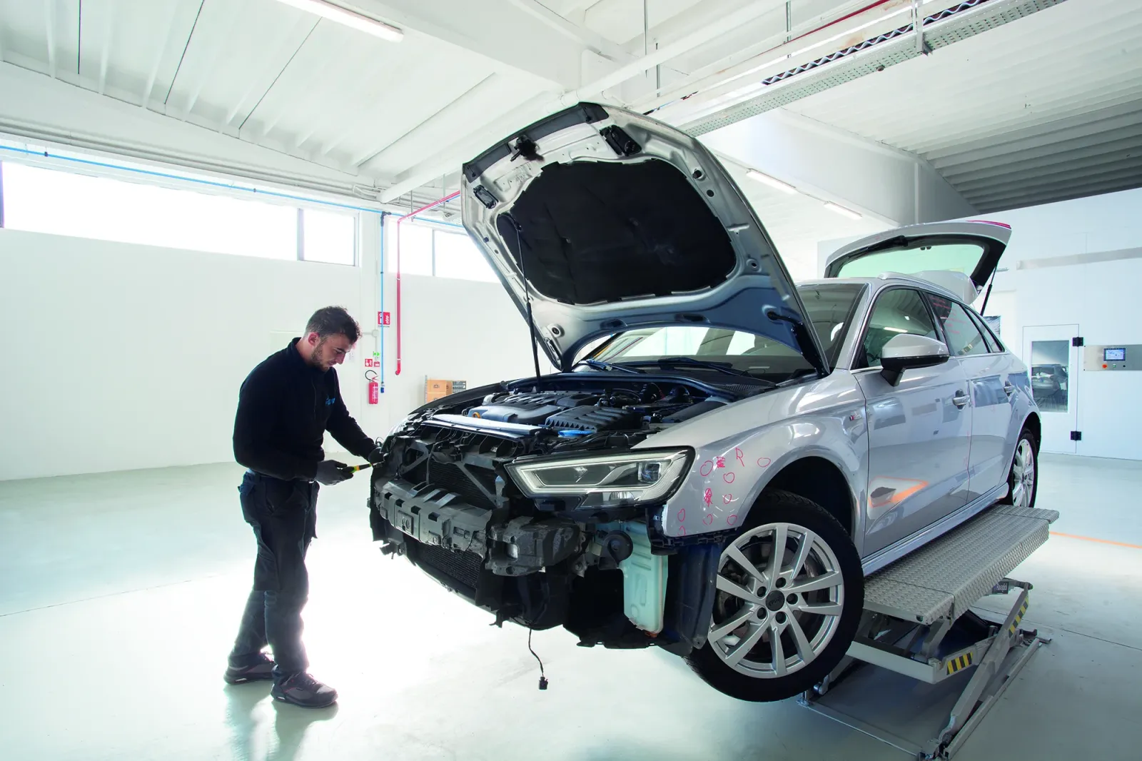 Un meccanico in officina esamina un'auto sollevata con cofano e paraurti aperti per la riparazione della carrozzeria.