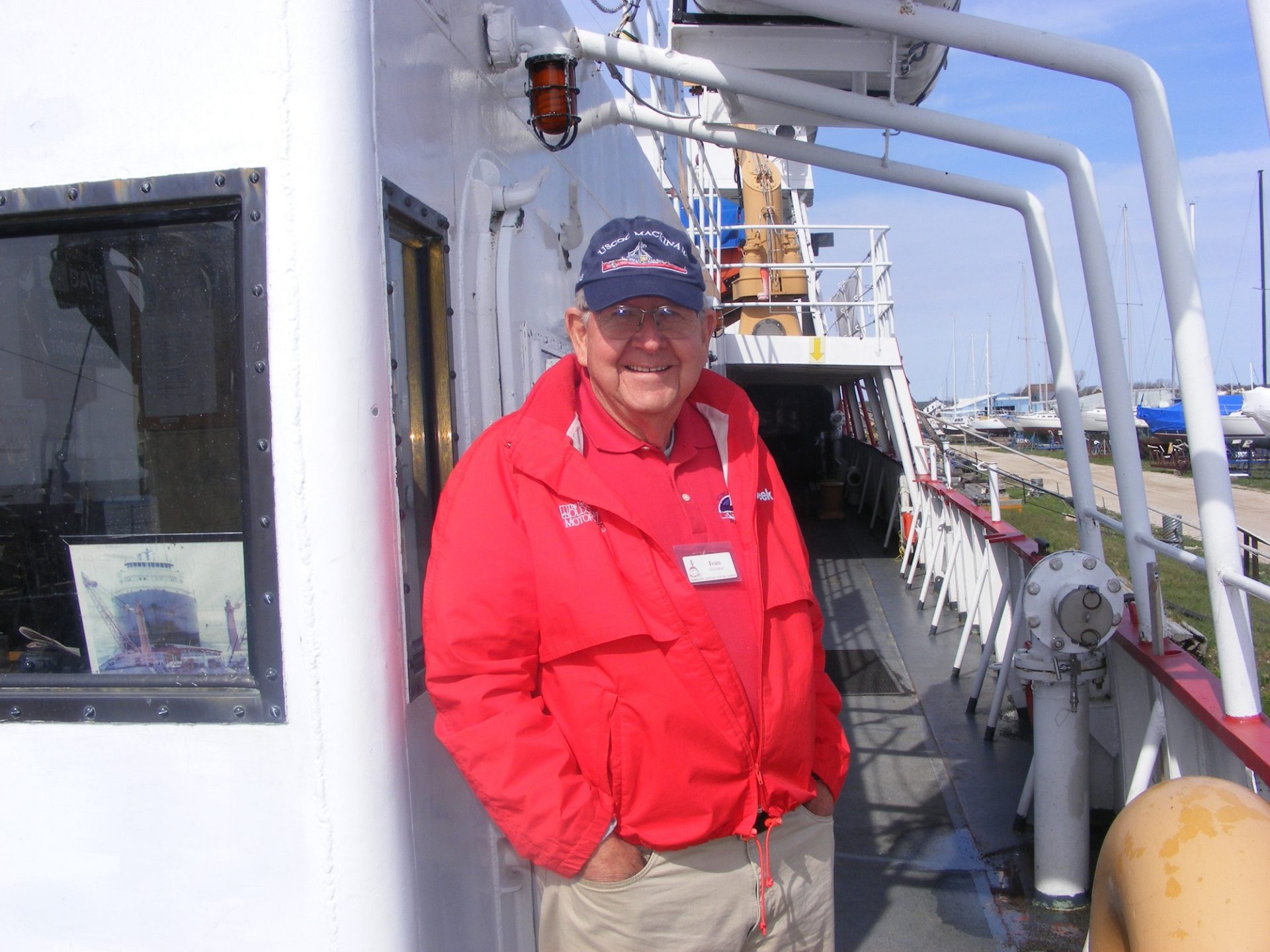 Smiling male volunteer in a red jacket and USCGC Mackinaw cap stands on the deck of the museum ship wearing a name badge.