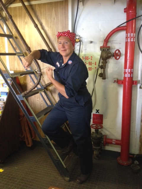 Woman dressed as Rosie the Riveter in blue coveralls and red polka-dot bandana poses on a ladder inside the USCGC Mackinaw.