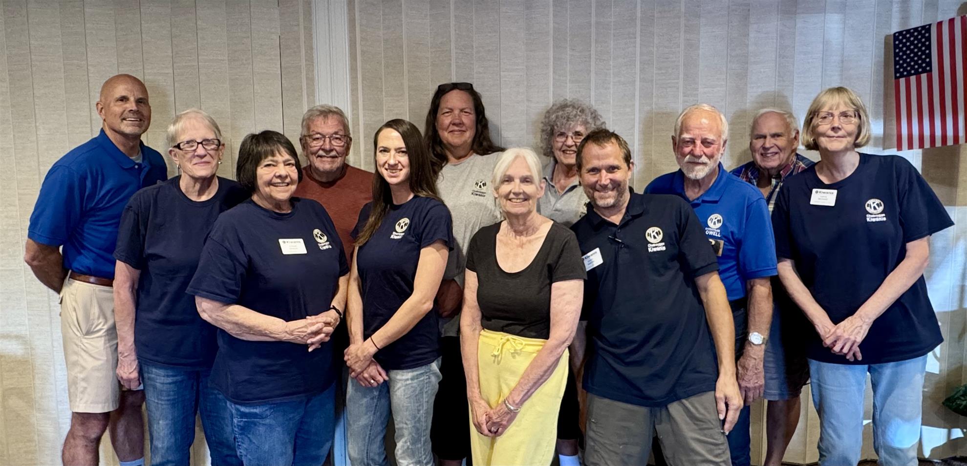 Group photo of Cheboygan Kiwanis Club members wearing navy Kiwanis shirts, posed indoors with an American flag in the background.
