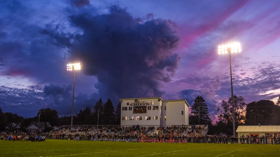 Cheboygan Chiefs football stadium at dusk, packed with spectators under stadium lights with dramatic purple and pink cloudy sky.