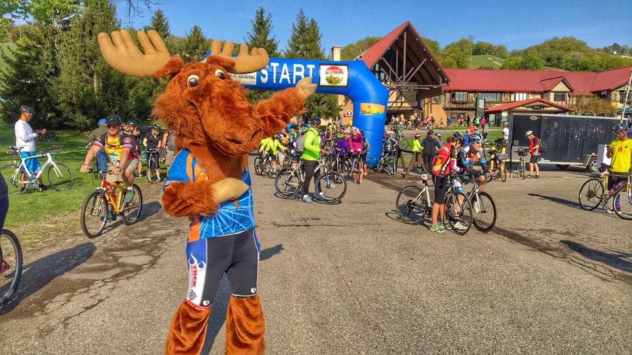 Zoo-de-Mack bike ride mascot moose at the START arch with cyclists gathered outdoors.