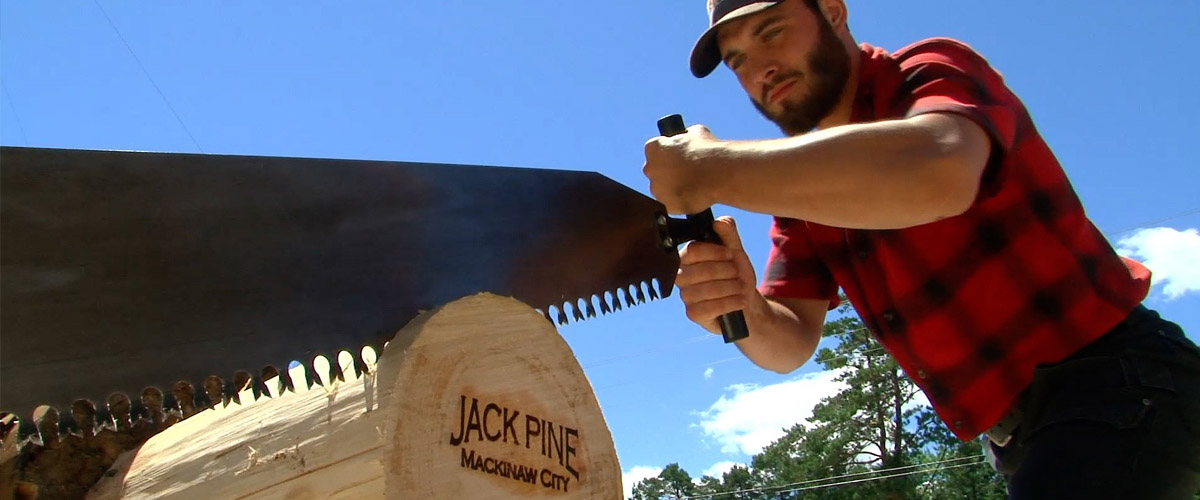 Jack Pine Lumberjack Shows in Mackinaw City — performer in red flannel crosscut-saws a log branded with the Jack Pine Mackinaw City logo.