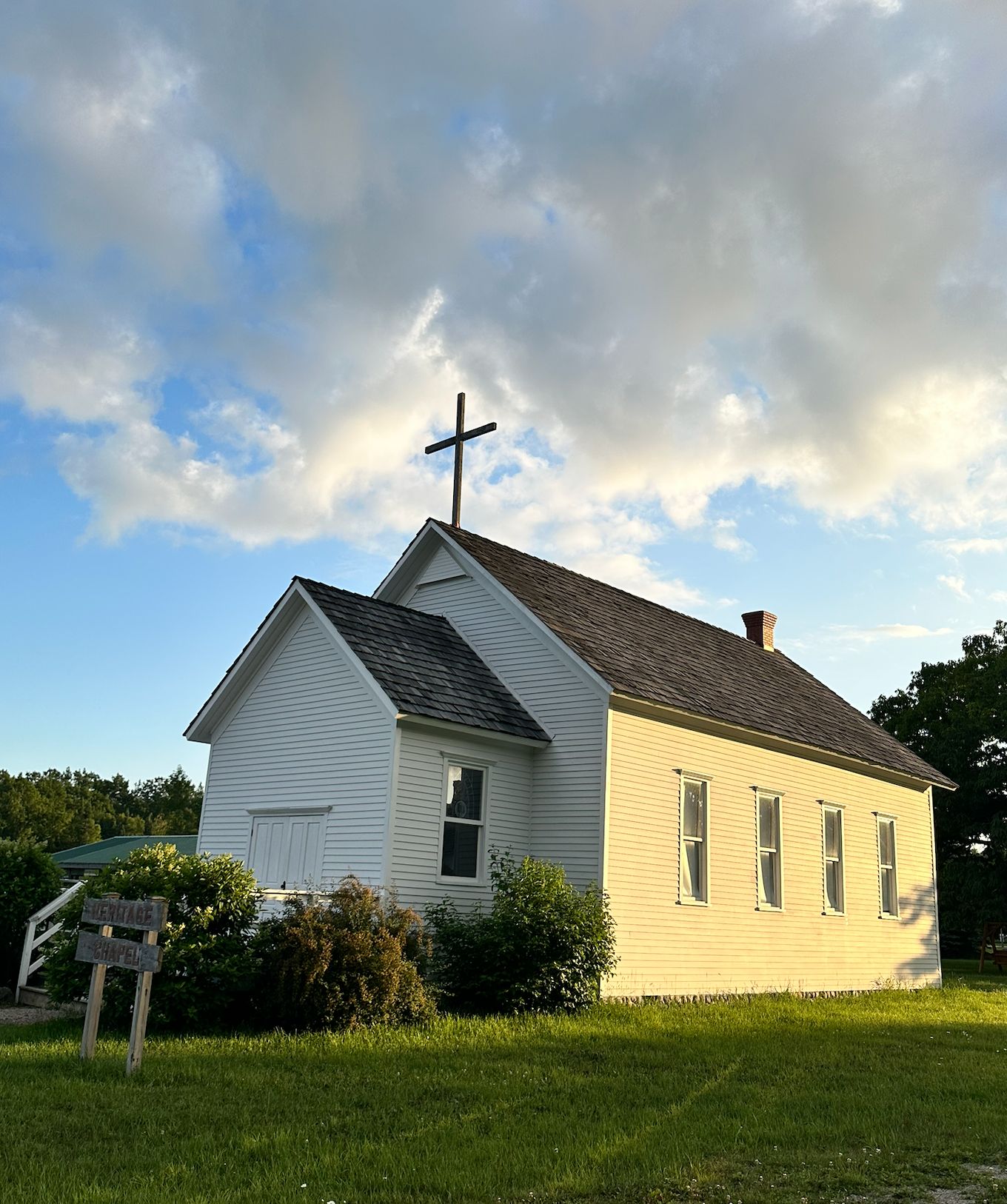 Heritage Chapel, a white building with a cross on top, stands surrounded by green grass and bushes under a partly cloudy sky.