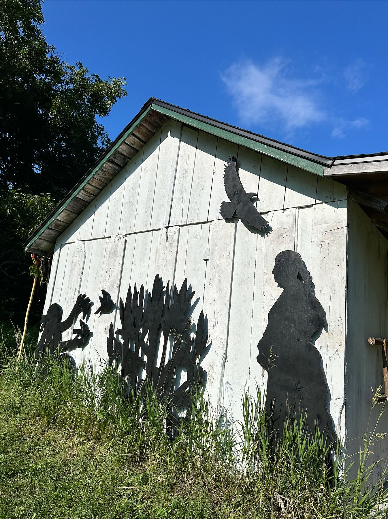 Silhouette cutouts of a woman, children, corn stalks, and birds decorate the side of a white building at Heritage Village, depicting the Anishinaabe Settlement.