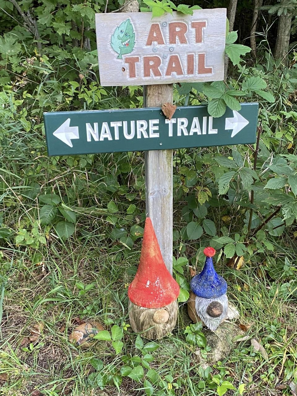 Wooden sign reading 'Art Trail' above a green 'Nature Trail' directional sign, with two hand-crafted gnome figures at the base, surrounded by lush greenery.