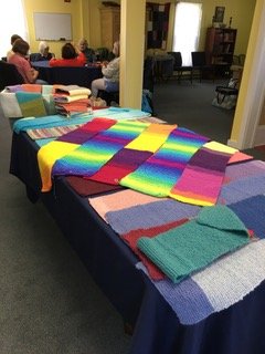 Colorful knitted and crocheted blankets displayed on a table at a church gathering, with participants seated in the background.