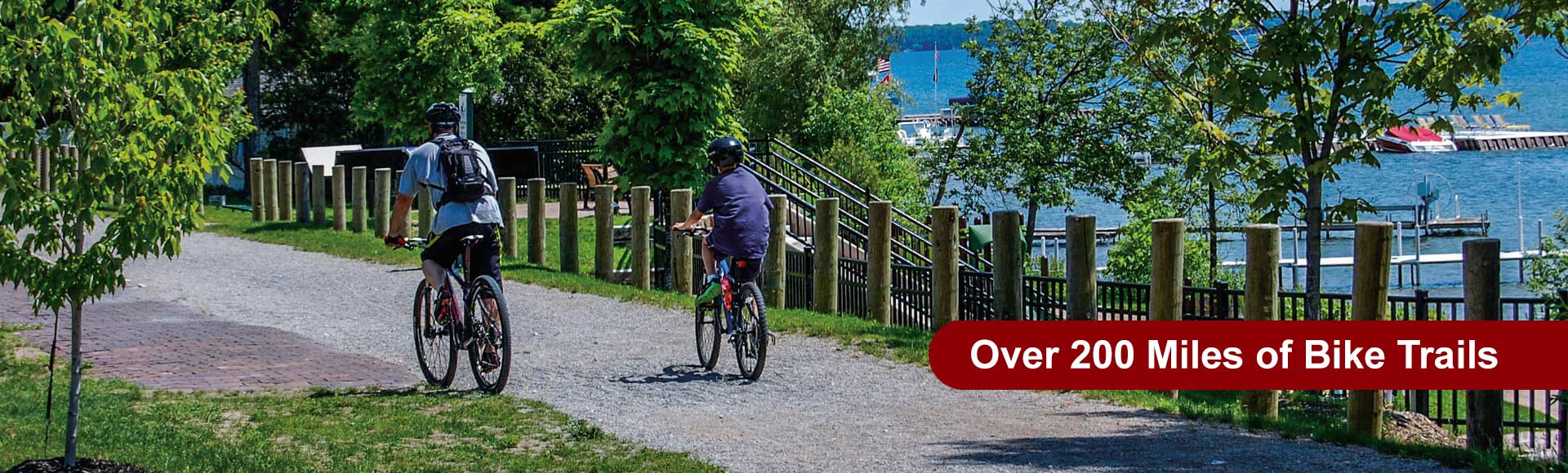 Two cyclists ride helmeted on a gravel trail beside a waterway in Cheboygan. Text reads: Over 200 Miles of Bike Trails.