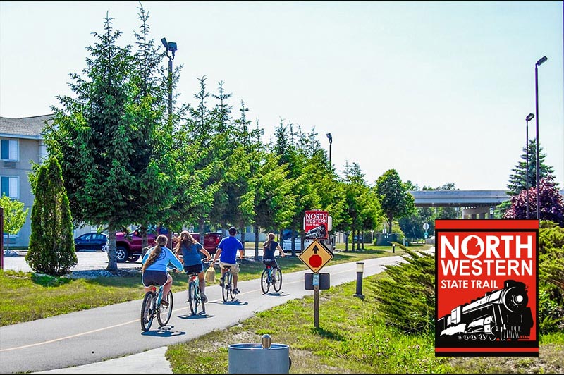 Group of cyclists ride on the paved North Western State Trail on a sunny day. North Western State Trail logo visible on signage.