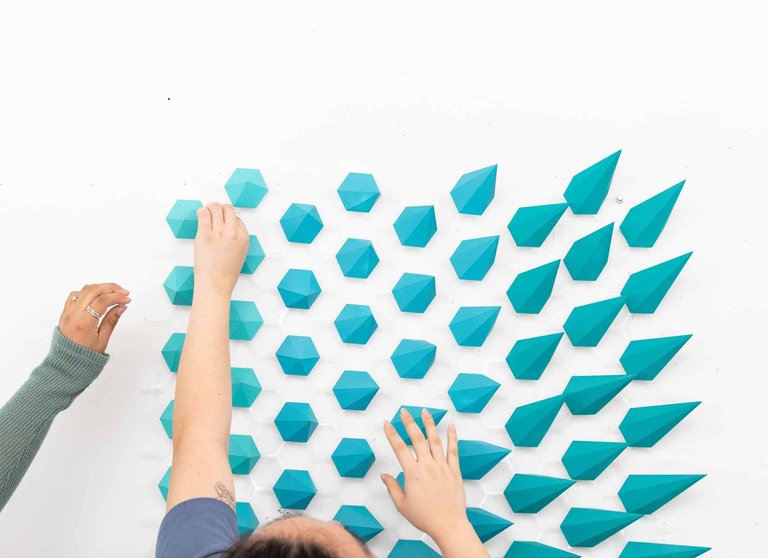 Two students install a series of teal, geometric pieces on a white wall.