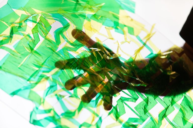 Abstract close-up photograph of translucent green and yellow tissue paper or cellophane fragments, with a silhouetted hand visible through the layers. The pieces create a dynamic, overlapping pattern with varying shades of emerald and chartreuse against a bright white background. The materials appear delicate and light-catching, creating an ethereal effect with their transparent nature. The composition suggests movement and lightness, with the fragments appearing to float or cascade across the frame.